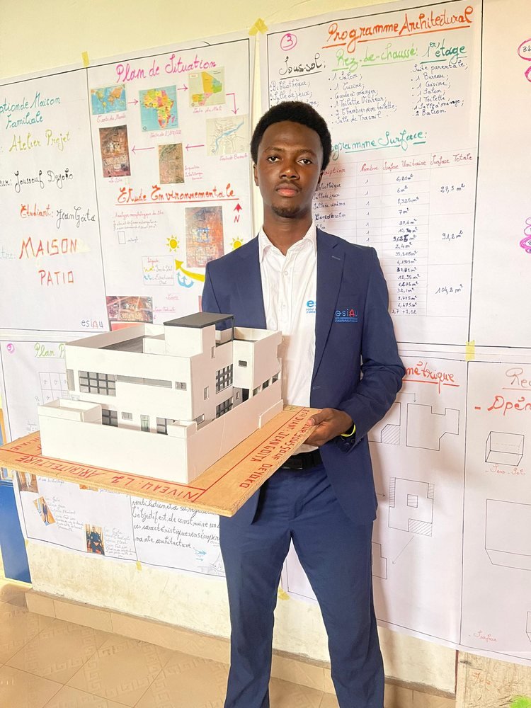 Un étudiant en costume présente la maquette d'une villa blanche devant ses planches